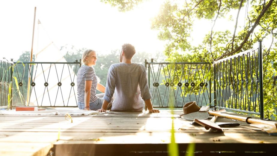 A couple sitting on an unfinished deck frame beside a jetty and a sailing boat on the water