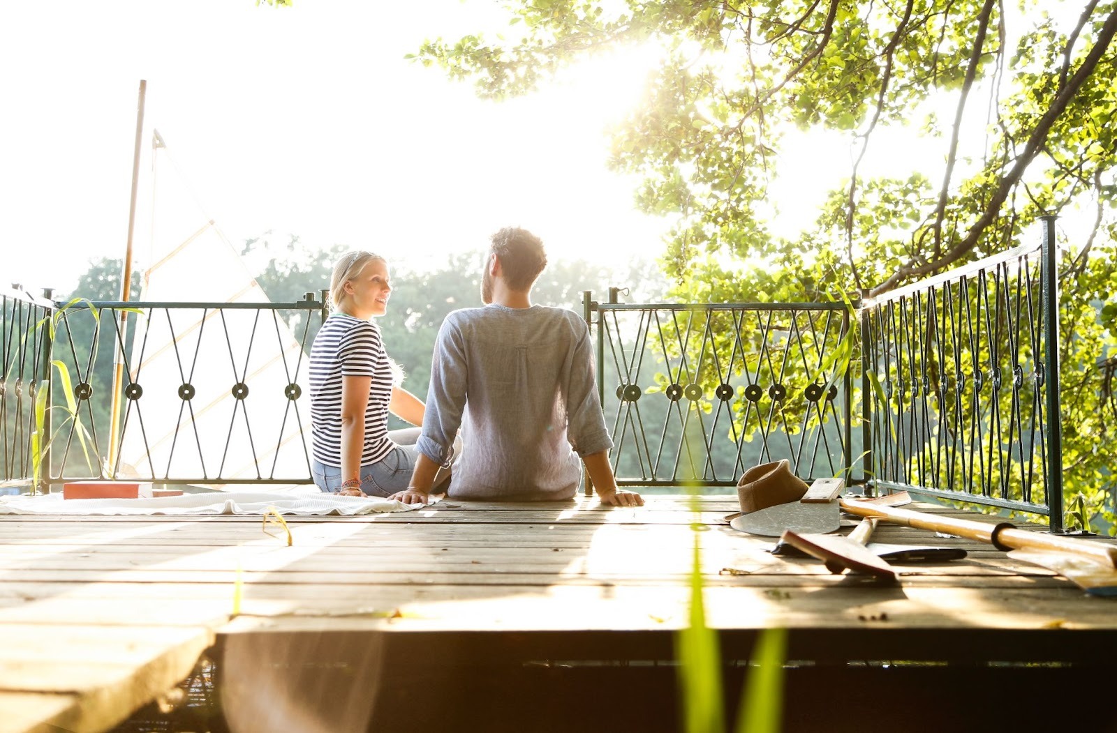 A couple sitting on an unfinished deck frame beside a jetty and a sailing boat on the water