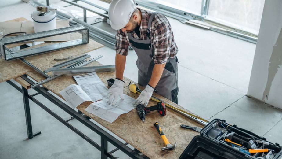 A general contractor reviewing construction blueprints on a worktable at a job site, surrounded by tools and building materials