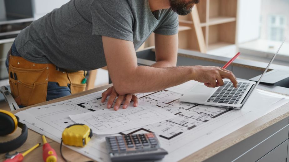 A male homeowner reviewing kitchen remodel prices on a laptop with blueprints, calculator, measuring tape, and renovation tools on the counter
