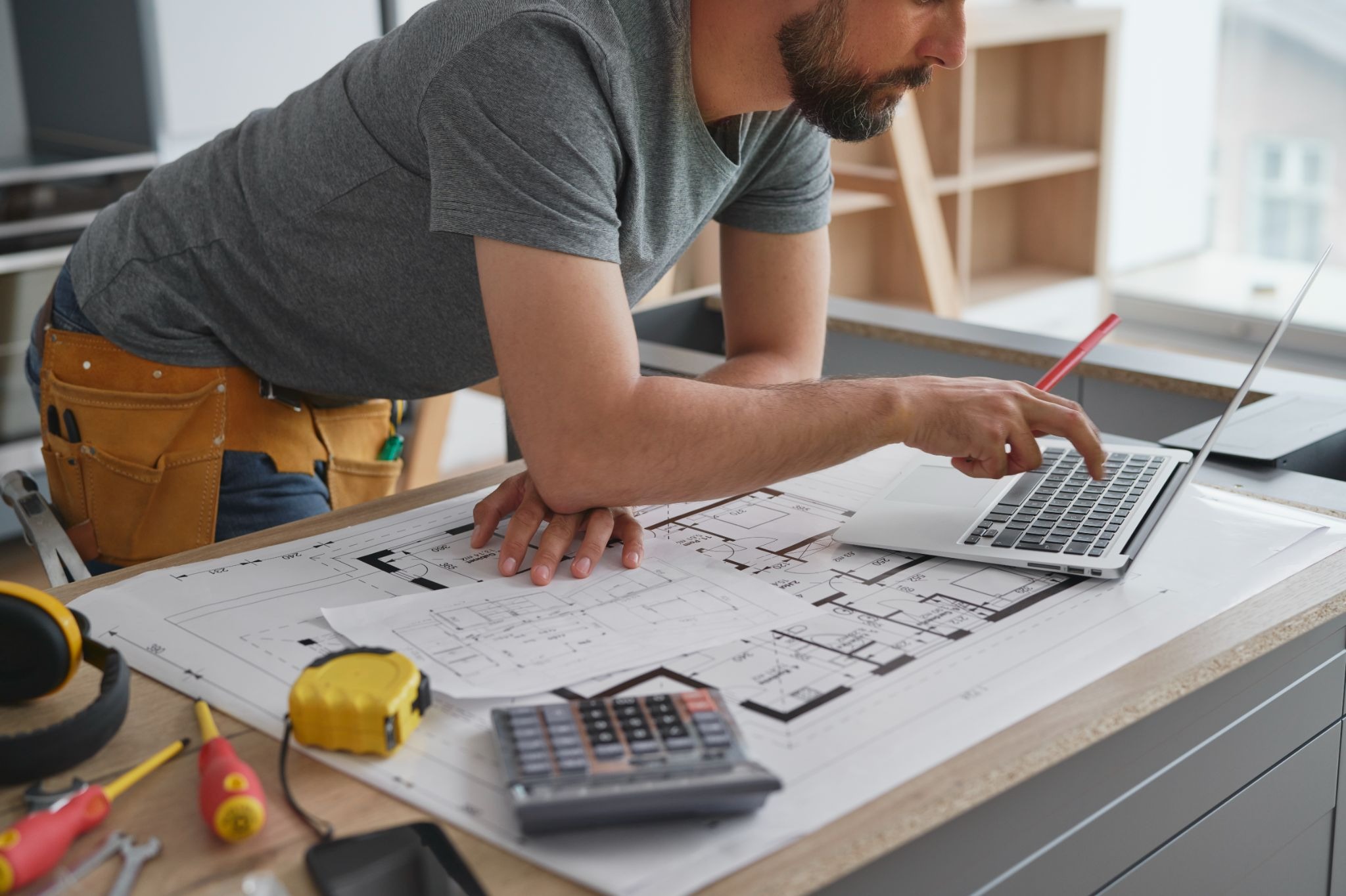 A male homeowner reviewing kitchen remodel prices on a laptop with blueprints, calculator, measuring tape, and renovation tools on the counter