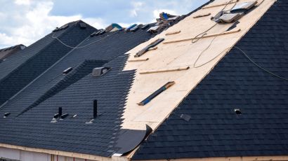 Residential roof under construction with dark shingles on one side and exposed plywood on the other, scattered boards and construction materials.