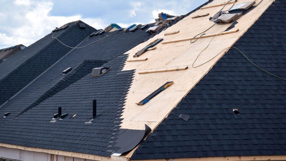 Residential roof under construction with dark shingles on one side and exposed plywood on the other, scattered boards and construction materials.