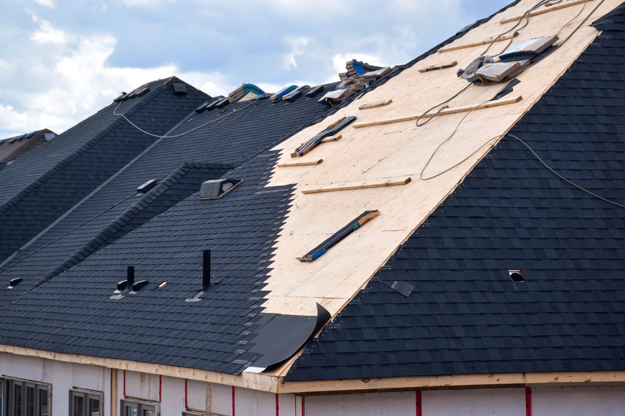 Residential roof under construction with dark shingles on one side and exposed plywood on the other, scattered boards and construction materials.